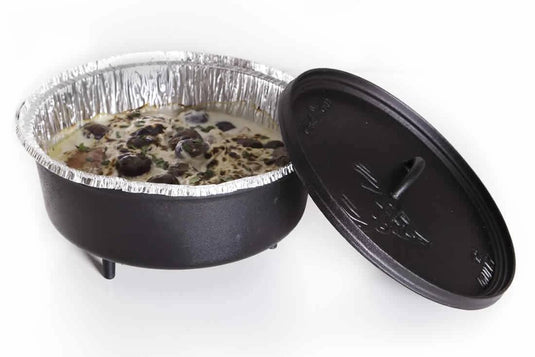 A black cast iron Dutch oven holds a foil-lined dish, possibly using Camp Chef Disposable Dutch Oven Liners, topped with black olives. The lid rests beside the pot on a white background—ideal for Dutch oven cooking.