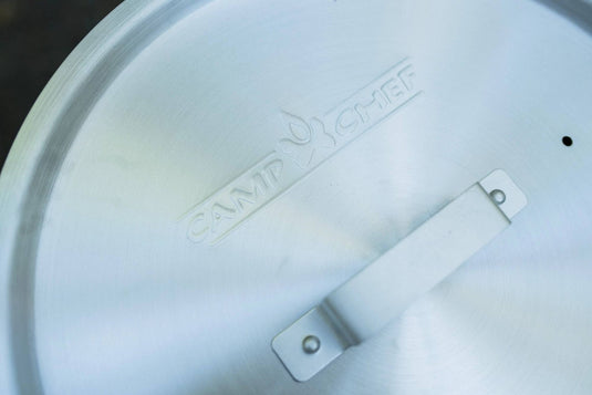 A close-up of the Camp Chef logo embossed on a metal lid with a handle, resting on the Aluminum Cooker Pot by Camp Chef, with a blurred background.