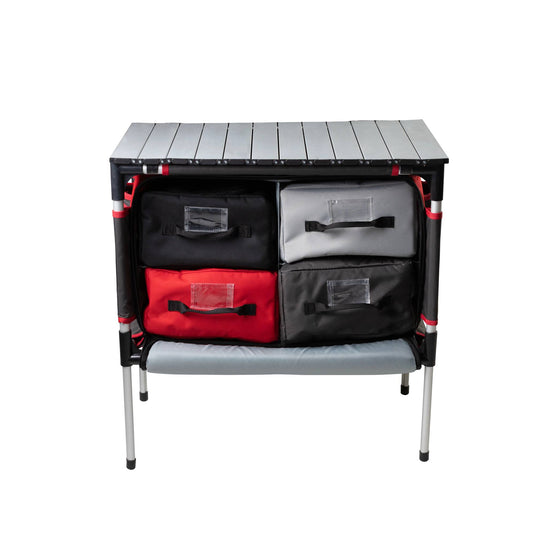The Camp Chef Sherpa Table & Organizer features a gray tabletop, four storage bins in black, gray, and red, a lower shelf, and silver legs—ideal as an outdoor cooking station or camp table—shown against a white background.