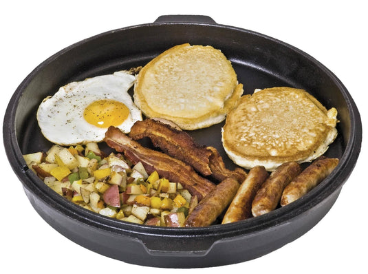 A Camp Chef Deluxe Dutch Oven filled with a breakfast meal: a sunny-side-up egg, two pancakes, crispy bacon strips, sausage links, and diced potatoes with peppers.