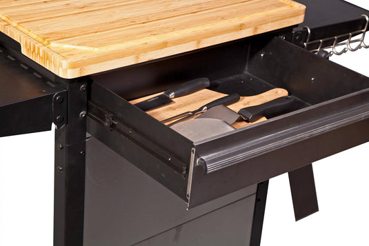 A close-up of the Camp Chef Patio Cart shows an open drawer with several kitchen knives beneath a wooden cutting board on top.