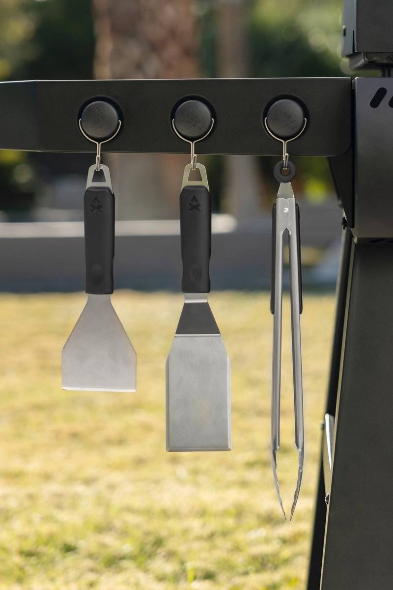 Load image into Gallery viewer, Three Camp Chef Magnetic Tool Holders (3 Pack) secure grilling tools—spatula, scraper, and tongs—to the grill’s side, with blurred grass and trees in the background.
