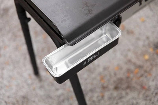 A close-up of Camp Chef Disposable Grease Cup Liners (5-Pk) in a rectangular metal drip tray under a black outdoor grill, positioned to catch grease or drippings. The gray ground beneath features scattered leaves.
