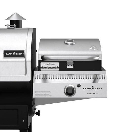 Close-up of a Camp Chef Sidekicks stainless steel accessory mounted on a larger grill, featuring control knobs, a closed lid with handle, and a temperature gauge for precise propane burner control.