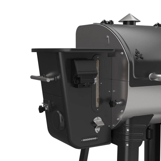 Close-up of the Camp Chef Woodwind 24 pellet grill in black and silver, highlighting the hopper with visible pellets and the WIFI controller. The unit includes a sturdy handle and multiple control knobs for easy operation.