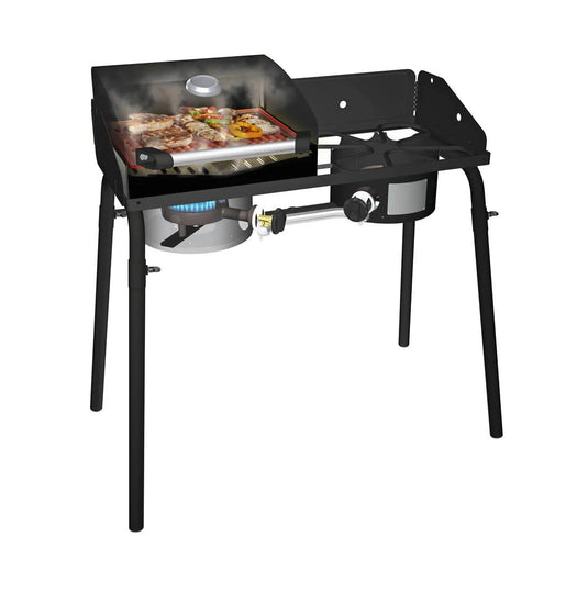 The Camp Chef BBQ Grill Boxes 14, a black propane grill with a griddle top and cast iron surface, is shown cooking meat and veggies. It has two burners (one in use), smoke rising, sizzling food, side shelf, and sturdy legs for support.