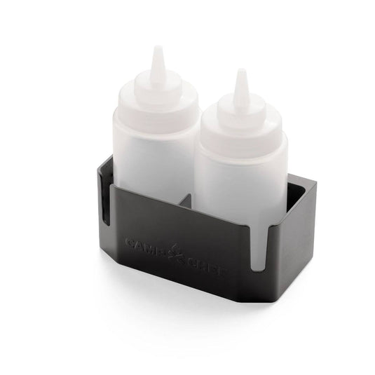 Two clear squeeze bottles with white caps sit upright in a black Bottle Caddy bottle holder by Camp Chef, embossed with the brand name on the side. The holder features an open front and is set against a white background.