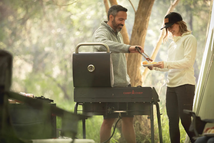 Two people grilling outdoors in a forest setting with a Camp Chef grill.